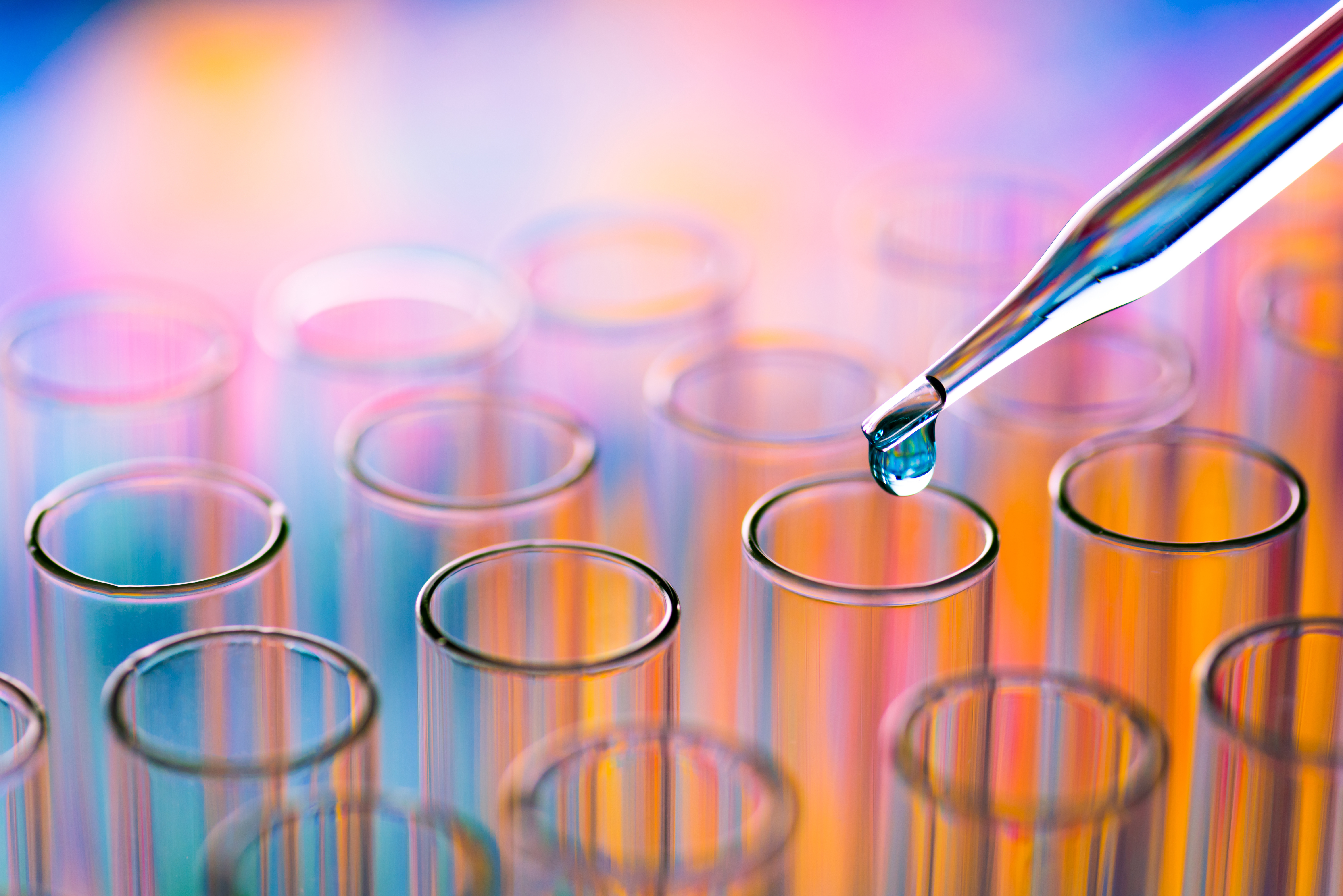 A pipette releases a water droplet into a test tube among an array of other colorful test tubes.